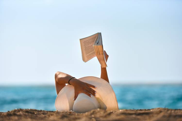 A person wearing a white hat and white nail polish on the beach reading a book